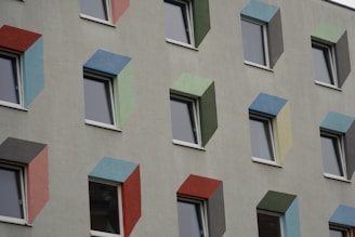 A building facade with multiple windows, each surrounded by frames in various colors such as red, blue, green, and yellow. The windows are evenly spaced and the frames create a geometric pattern on the otherwise plain gray wall.