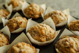 A batch of freshly baked muffins are arranged closely together in white paper cups. The muffins appear golden-brown with a crumbly textured top, suggesting a delicious and homemade quality.