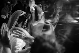 A black and white photograph captures a wedding celebration with a bride and groom surrounded by people clapping and cheering. The bride is wearing a veil and the groom is dressed in a suit. The image conveys a sense of movement and excitement through blurred hands and faces.