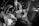 A black and white photograph captures a wedding celebration with a bride and groom surrounded by people clapping and cheering. The bride is wearing a veil and the groom is dressed in a suit. The image conveys a sense of movement and excitement through blurred hands and faces.