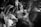 A black and white photograph captures a wedding celebration with a bride and groom surrounded by people clapping and cheering. The bride is wearing a veil and the groom is dressed in a suit. The image conveys a sense of movement and excitement through blurred hands and faces.