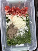A clear plastic container holds a variety of chopped ingredients, including red tomatoes, white onions, green herbs, ground pepper, and salt. The ingredients are neatly arranged in sections, creating a colorful display of fresh produce and seasonings.