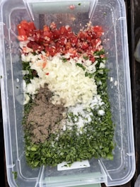 A clear plastic container holds a variety of chopped ingredients, including red tomatoes, white onions, green herbs, ground pepper, and salt. The ingredients are neatly arranged in sections, creating a colorful display of fresh produce and seasonings.