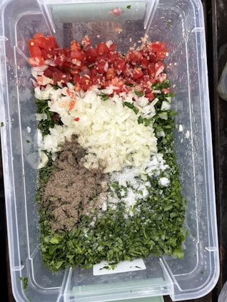 A clear plastic container holds a variety of chopped ingredients, including red tomatoes, white onions, green herbs, ground pepper, and salt. The ingredients are neatly arranged in sections, creating a colorful display of fresh produce and seasonings.