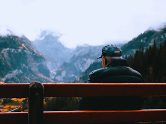 A quiet moment of reflection, a man sitting on a weathered bench overlooking vast mountains under a darkening sky.