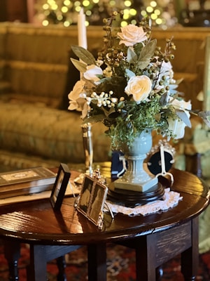 A bouquet of white roses and greenery is elegantly arranged in a vase on a polished wooden table. Surrounding the flowers are books, framed photographs, a doily, and a candle. The background features a sofa with subtle, warm lighting and a bokeh effect from decorative lights.