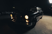 A vintage car with its headlights on is parked in a dimly lit garage. The vehicle is mostly in shadow, with only the front part partially illuminated, highlighting its classic design. Other vintage cars can be faintly seen in the background, adding to the nostalgic atmosphere.