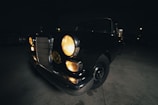 A vintage car is parked indoors in a dimly lit environment. The headlights are on, casting a warm glow. The car features a classic design with round headlights and a notable front grille.