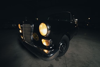 A vintage car is parked indoors in a dimly lit environment. The headlights are on, casting a warm glow. The car features a classic design with round headlights and a notable front grille.