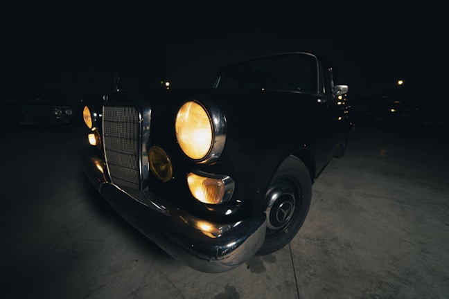 A vintage car is parked indoors in a dimly lit environment. The headlights are on, casting a warm glow. The car features a classic design with round headlights and a notable front grille.