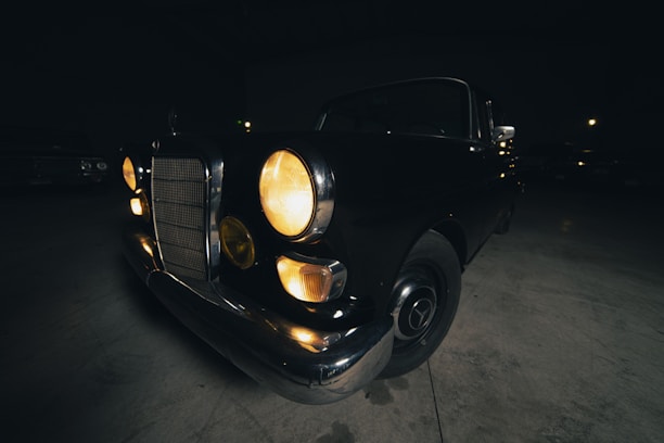 A vintage car is parked indoors in a dimly lit environment. The headlights are on, casting a warm glow. The car features a classic design with round headlights and a notable front grille.