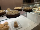 A bakery display case containing several pastries and cakes. Three large cakes with lattice tops are prominently featured on a glass shelf, with one cake partially sliced. Below on a white plate, there are cream-filled pastries and a croissant.