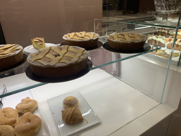 A bakery display case containing several pastries and cakes. Three large cakes with lattice tops are prominently featured on a glass shelf, with one cake partially sliced. Below on a white plate, there are cream-filled pastries and a croissant.