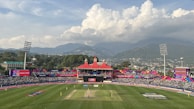 A cricket stadium filled with spectators against a backdrop of mountains and a cloudy sky. The field is vibrant green, and players can be seen on the pitch with a colorful stadium decor in the background. The stands are crowded with people, and large floodlights tower over the scene.