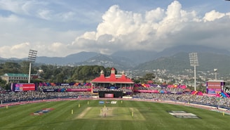 A cricket stadium filled with spectators against a backdrop of mountains and a cloudy sky. The field is vibrant green, and players can be seen on the pitch with a colorful stadium decor in the background. The stands are crowded with people, and large floodlights tower over the scene.