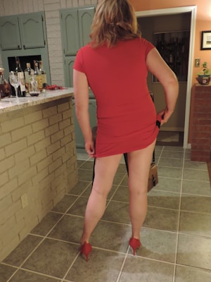 A person in a red dress and matching red high heels stands in a kitchen with their back to the camera. Various bottles and a wine glass are placed on a tiled countertop. The kitchen features teal-colored cabinets and beige brick walls.