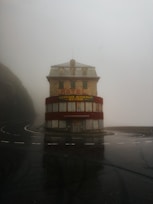 A solitary, weathered hotel building stands at a curve in a wet, foggy road. The structure is multi-storied with red accents and signage in German. The area is enveloped in mist, creating an eerie, isolated atmosphere.