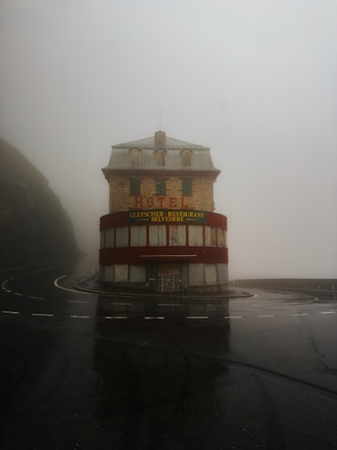 A solitary, weathered hotel building stands at a curve in a wet, foggy road. The structure is multi-storied with red accents and signage in German. The area is enveloped in mist, creating an eerie, isolated atmosphere.