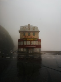 A solitary, weathered hotel building stands at a curve in a wet, foggy road. The structure is multi-storied with red accents and signage in German. The area is enveloped in mist, creating an eerie, isolated atmosphere.