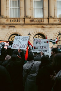 A group of people gathered in front of a large building, holding signs with messages advocating against genocide and calling for a ceasefire. Some Palestinian flags are visible among the crowd.