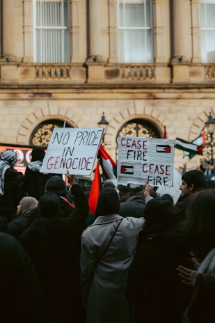 A group of people gathered in front of a large building, holding signs with messages advocating against genocide and calling for a ceasefire. Some Palestinian flags are visible among the crowd.