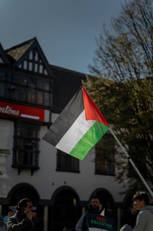 A Palestinian flag is prominently displayed on a flagpole against the backdrop of a building with traditional architecture and a clear blue sky. Several individuals stand nearby, one holding a sign that reads 'Free Palestine.' Trees with sparse leaves are visible on the side.