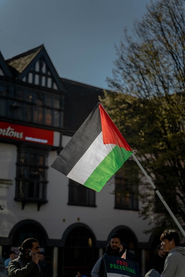 A Palestinian flag is prominently displayed on a flagpole against the backdrop of a building with traditional architecture and a clear blue sky. Several individuals stand nearby, one holding a sign that reads 'Free Palestine.' Trees with sparse leaves are visible on the side.