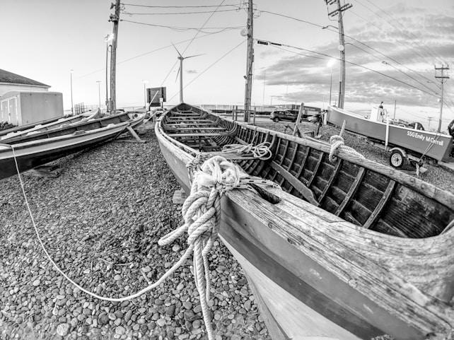 A black and white image features a wooden boat on a pebbled area, with ropes coiled on and around it. Another boat is visible in the background alongside a utility pole and a wind turbine. The sky is cloudy, and various structures and vehicles can be seen in the distance.