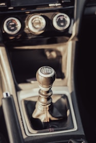 A close-up view of a manual gear shift lever inside a vehicle, surrounded by a dark console with three circular climate control knobs above it. The leather-wrapped gear knob displays shift pattern markings.