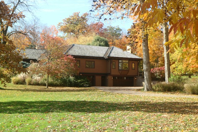 A house surrounded by a lush garden and abundant trees displaying autumn foliage, with shades of orange, yellow, and red. The grass is mostly green with scattered fallen leaves.