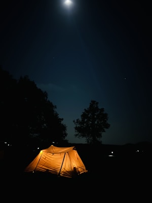A glowing tent is illuminated from within under a starry night sky, with the bright moon casting a gentle light over the scene. Surrounding the tent are silhouetted trees and a serene outdoor setting, evoking a sense of tranquility and solitude.