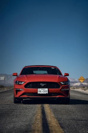 A red sports car is parked on a deserted road under a clear blue sky. The Mustang emblem is visible on the front grille, and the car has a Texas license plate. In the background, a yellow road sign is seen, and mountains are faintly visible.