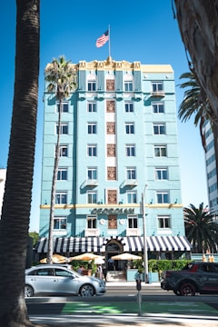 A multi-story building with an art deco design, painted in a light turquoise color. The building features a symmetrical facade with ornate decorations and numerous windows. At the top, an American flag is prominently displayed. A couple of palm trees are visible nearby, and there are parked cars along the street in front of the building.