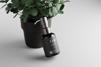 A black pump bottle labeled 'TINY' rests against a dark green potted plant with round leaves. The bottle has a metallic pump and is placed on a smooth, light-colored surface, creating a minimalist and elegant aesthetic.