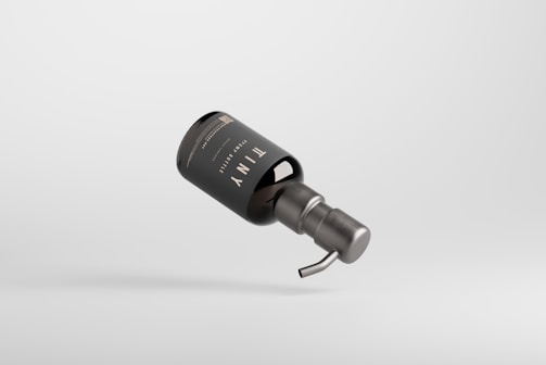 A sleek, dark-colored pump bottle with minimalist labeling. The bottle appears to be floating against a plain, light gray background. The design is modern and clean, emphasizing a sophisticated aesthetic.