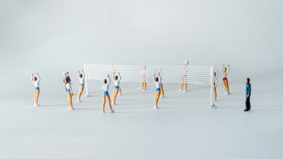 A scene of small figurines playing volleyball. The figures are dressed in athletic attire, with a volleyball net set up in the middle. There are several players on each side, poised as if in the middle of a game. One figure, dressed differently, appears to be observing the game.