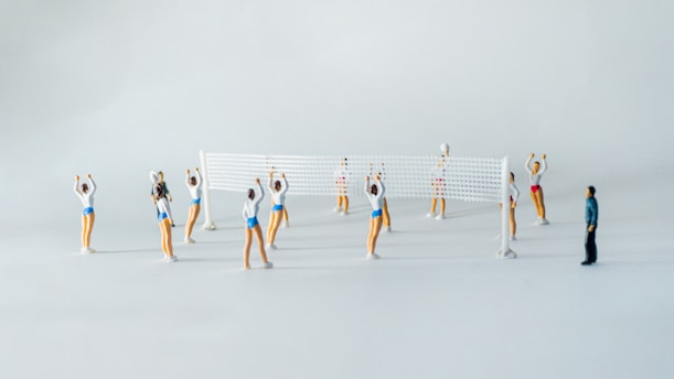 A scene of small figurines playing volleyball. The figures are dressed in athletic attire, with a volleyball net set up in the middle. There are several players on each side, poised as if in the middle of a game. One figure, dressed differently, appears to be observing the game.