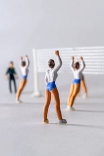Miniature figurines appear to be participating in a volleyball game. The figures wear white shirts and blue shorts, positioned around a white net. One figure stands apart, seemingly observing or officiating.