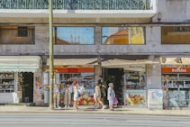 People are standing in a line in front of small shops under a concrete building. The shops feature signs with Portuguese words, suggesting a location in Portugal or a Portuguese-speaking region. There are multiple store fronts with windows reflecting nearby architecture.