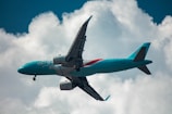 A teal-colored passenger airplane is flying against a backdrop of white clouds and a blue sky. The aircraft has distinct branding on its fuselage and is captured at an upward angle.