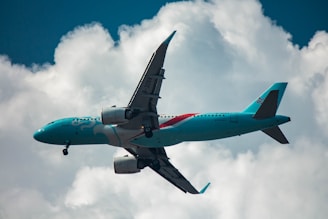 A teal-colored passenger airplane is flying against a backdrop of white clouds and a blue sky. The aircraft has distinct branding on its fuselage and is captured at an upward angle.
