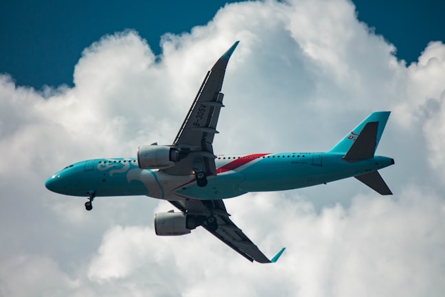 A teal-colored passenger airplane is flying against a backdrop of white clouds and a blue sky. The aircraft has distinct branding on its fuselage and is captured at an upward angle.
