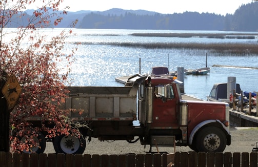 A red semi-truck is parked beside a lake with glistening water under the sunlight. The scene includes a few boats docked along the shore and a sign with a dog silhouette and the letters 'LAB' and 'XING'. A fence and a tree with red leaves are in the foreground, while mountains and a forested area can be seen in the background.