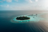 Aerial view of a small tropical island surrounded by vibrant turquoise waters. The island is lush with dense green vegetation and features a circular arrangement of overwater bungalows extending into the ocean, connected by a wooden pier. The vast expanse of the ocean stretches to the horizon under a partly cloudy sky.