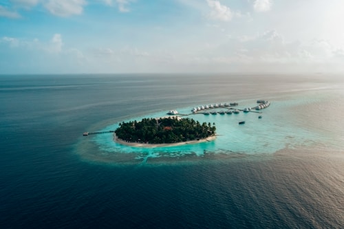 Aerial view of a small tropical island surrounded by vibrant turquoise waters. The island is lush with dense green vegetation and features a circular arrangement of overwater bungalows extending into the ocean, connected by a wooden pier. The vast expanse of the ocean stretches to the horizon under a partly cloudy sky.