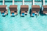 Aerial view of overwater bungalows with private pools, aligned on a turquoise sea. The structures feature brown roofs and are connected by wooden walkways, creating a symmetrical and serene vacation setting.