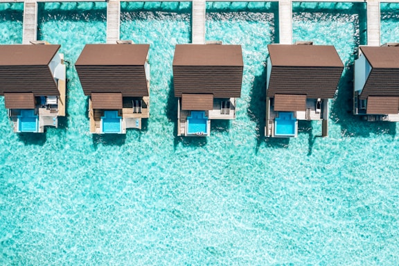 Aerial view of overwater bungalows with private pools, aligned on a turquoise sea. The structures feature brown roofs and are connected by wooden walkways, creating a symmetrical and serene vacation setting.