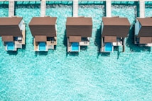 Aerial view of overwater bungalows with private pools, aligned on a turquoise sea. The structures feature brown roofs and are connected by wooden walkways, creating a symmetrical and serene vacation setting.