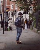 A person wearing a green jacket, jeans, and brown shoes is riding a skateboard on a city sidewalk. They have a dark backpack and are wearing headphones. The scene includes urban elements like traffic lights, street signs, and a building with red brick and white window frames. Several people are visible in the background, some wearing backpacks.