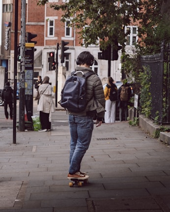 A person wearing a green jacket, jeans, and brown shoes is riding a skateboard on a city sidewalk. They have a dark backpack and are wearing headphones. The scene includes urban elements like traffic lights, street signs, and a building with red brick and white window frames. Several people are visible in the background, some wearing backpacks.